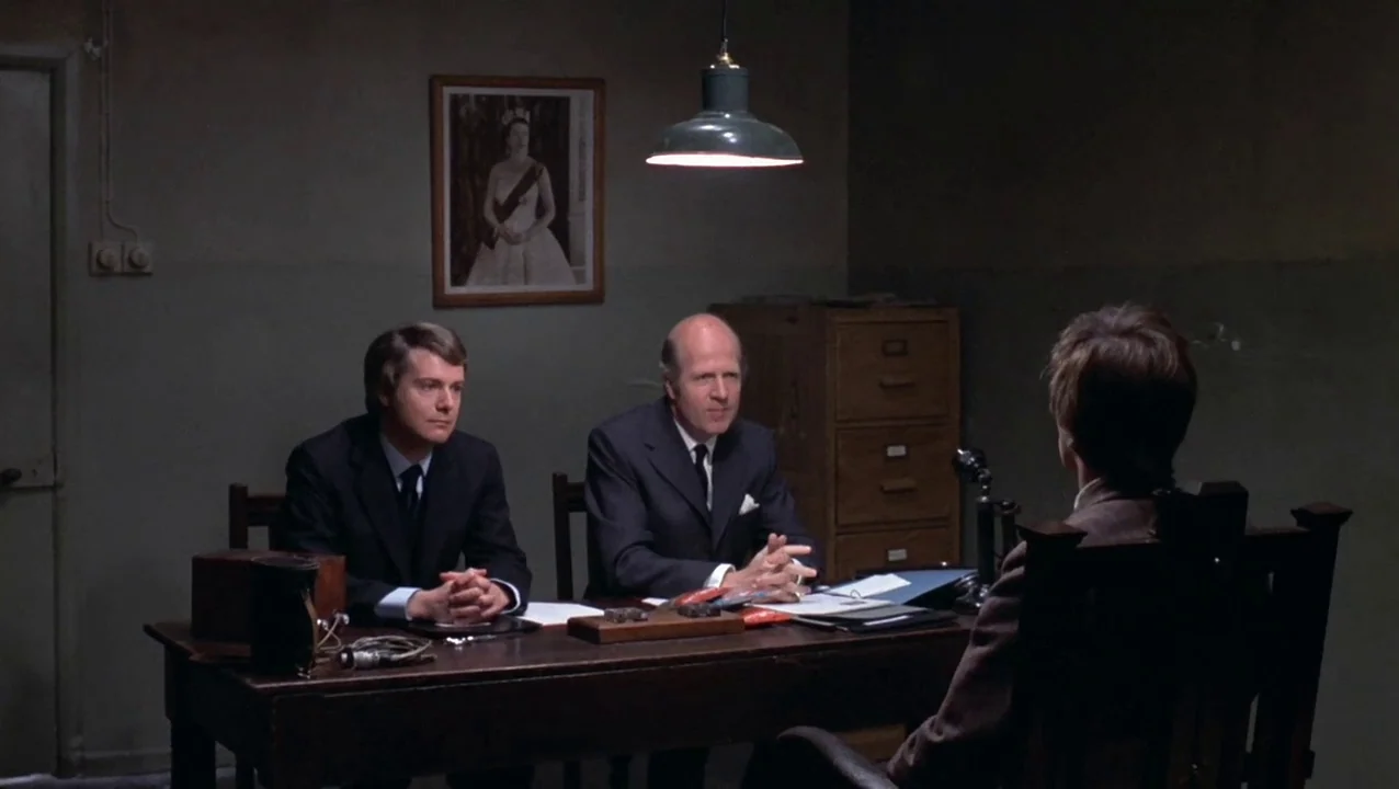 O Lucky Man! (1973) movie still: medium — A austere room features a dark wooden desk and a portrait on the wall. Two men in formal business suits…