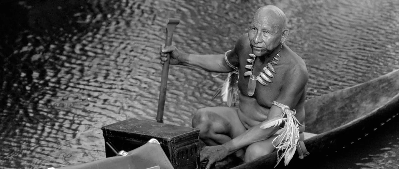 Embrace Of The Serpent (2015) movie still: medium — An elderly indigenous man sits inside a wooden canoe floating on dark water. He holds a paddle…
