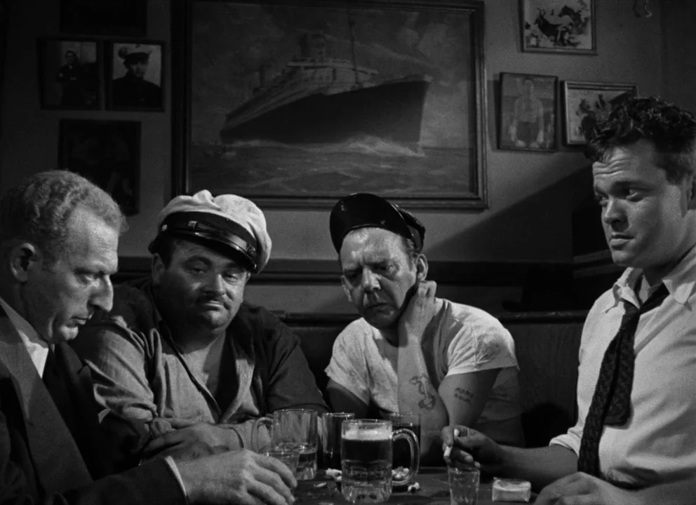 The Lady from Shanghai (1947) movie still: medium — A dimly lit 1940s tavern with nautical wall paintings and framed vintage photographs. Four men…