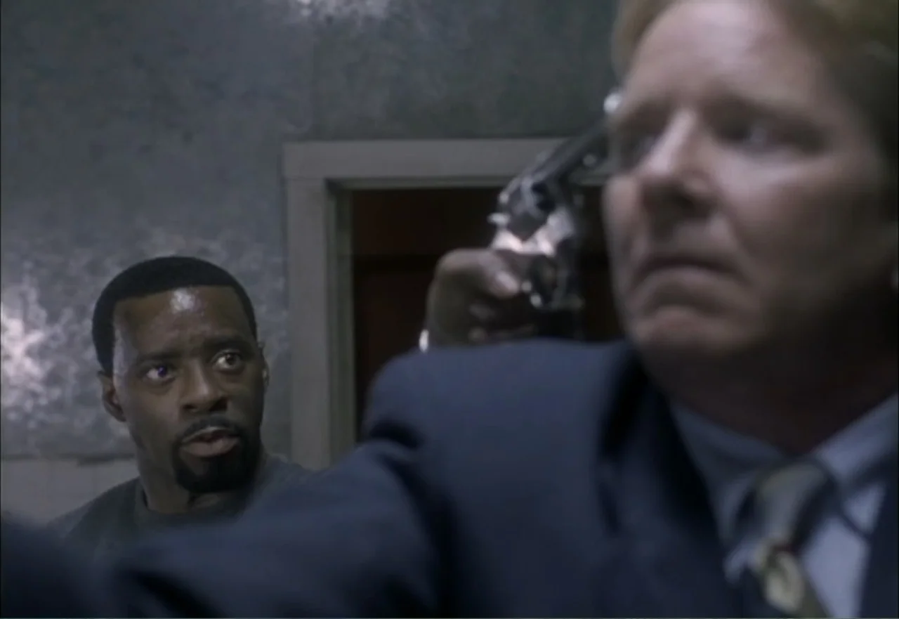 Ambushed (1998) movie still: medium — A dimly lit indoor corridor with plain walls and a door frame in the background. A man holds a pistol to the…