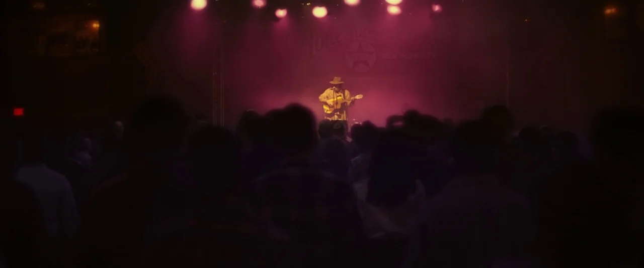 Blaze (2018) movie still: wide — A dark concert venue features a musician performing on a stage lit with purple light. A blurry crowd of silhouettes…