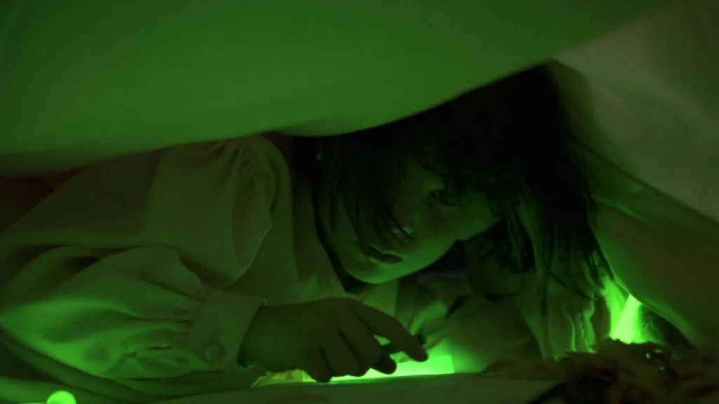 Cronos (1993) movie still: closeup — A child sits inside a makeshift fabric tent illuminated by a single green light. The soft fabric walls curve…