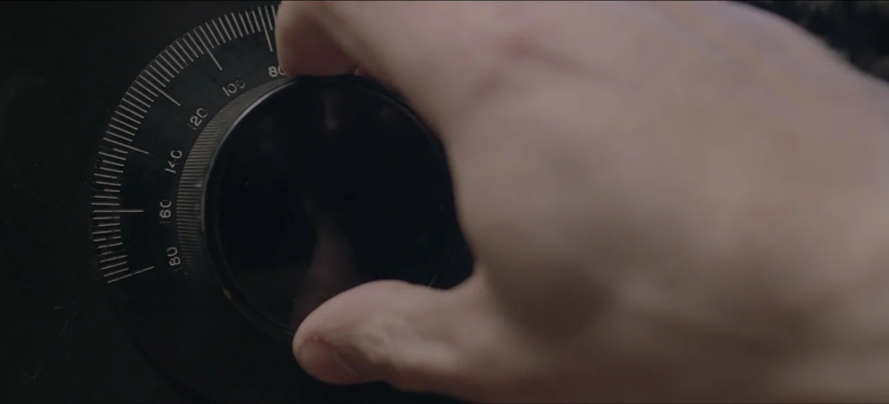 We Go In at Dawn (2020) movie still: closeup — A close-up view shows a metal dial on a black safe face. A hand rotates the dial slowly across the…