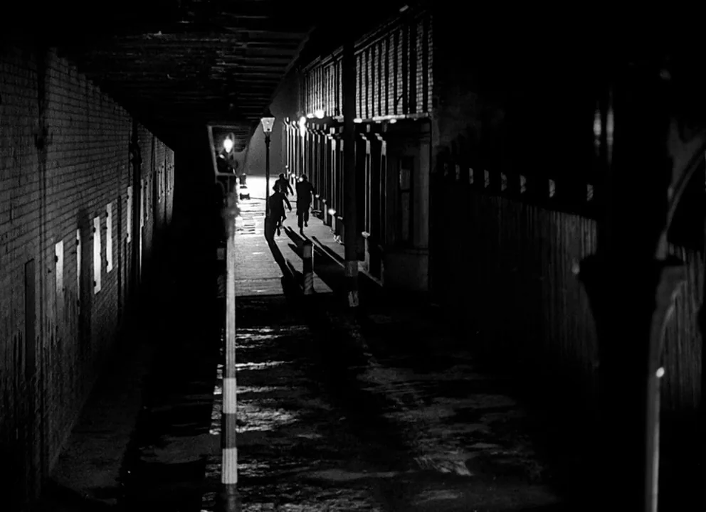 Odd Man Out (1947) movie still: wide — A narrow cobblestone urban alley at night features brick walls and overhead structures. Three figures in dark…