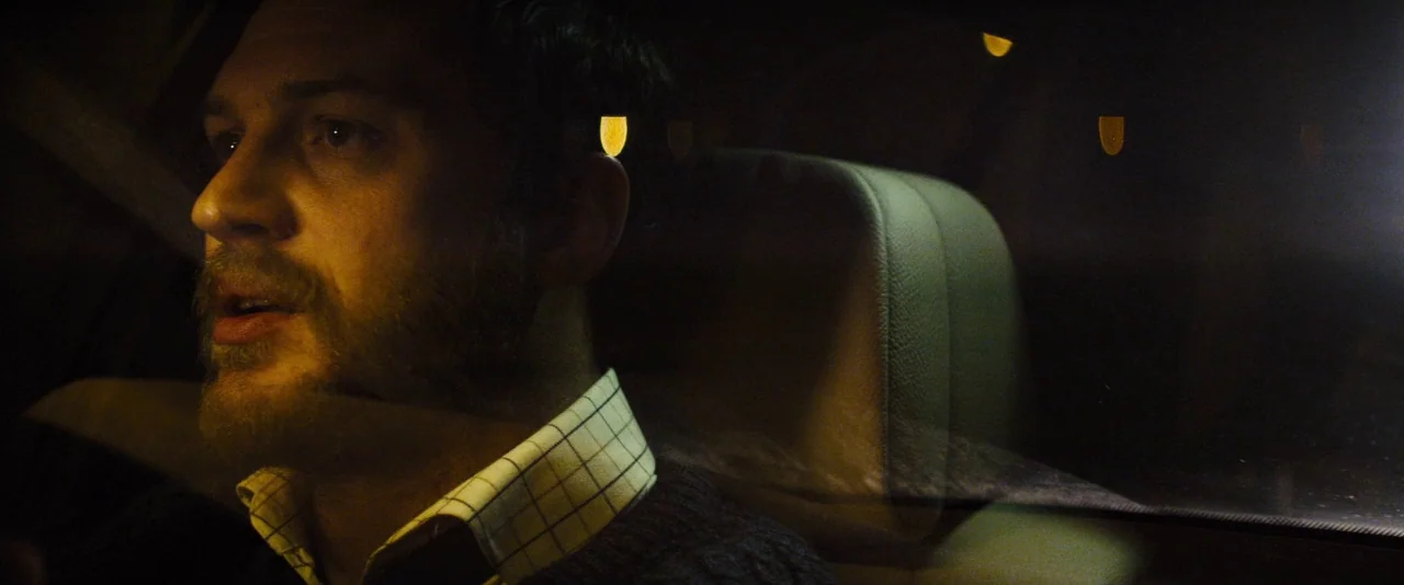 Locke (2013) movie still: closeup — The interior of a vehicle at night with blurred reflections on the window glass. A man sits in the driver seat…