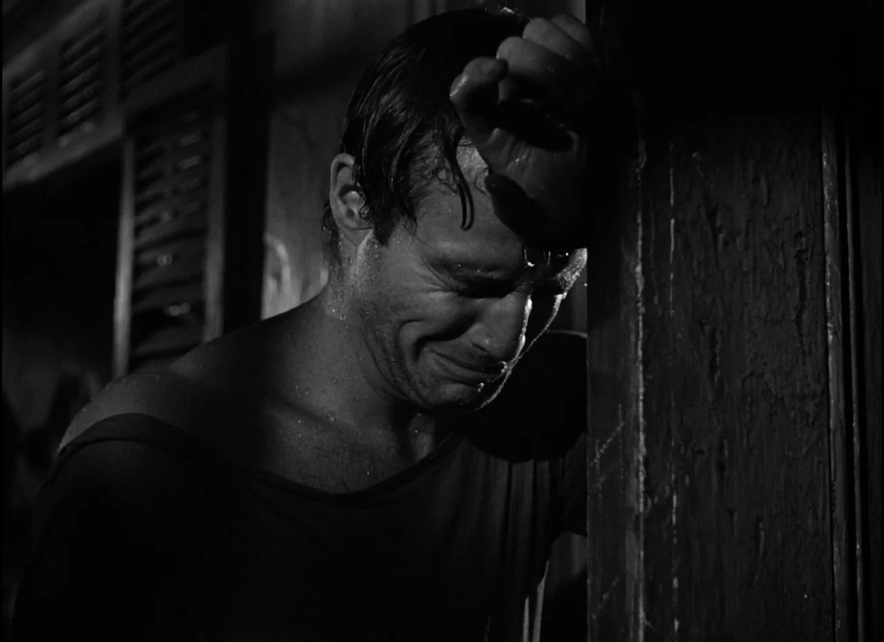 A Streetcar Named Desire (1951) movie still: closeup — A dimly lit interior space features a wooden doorframe and wall. A man rests his head against…