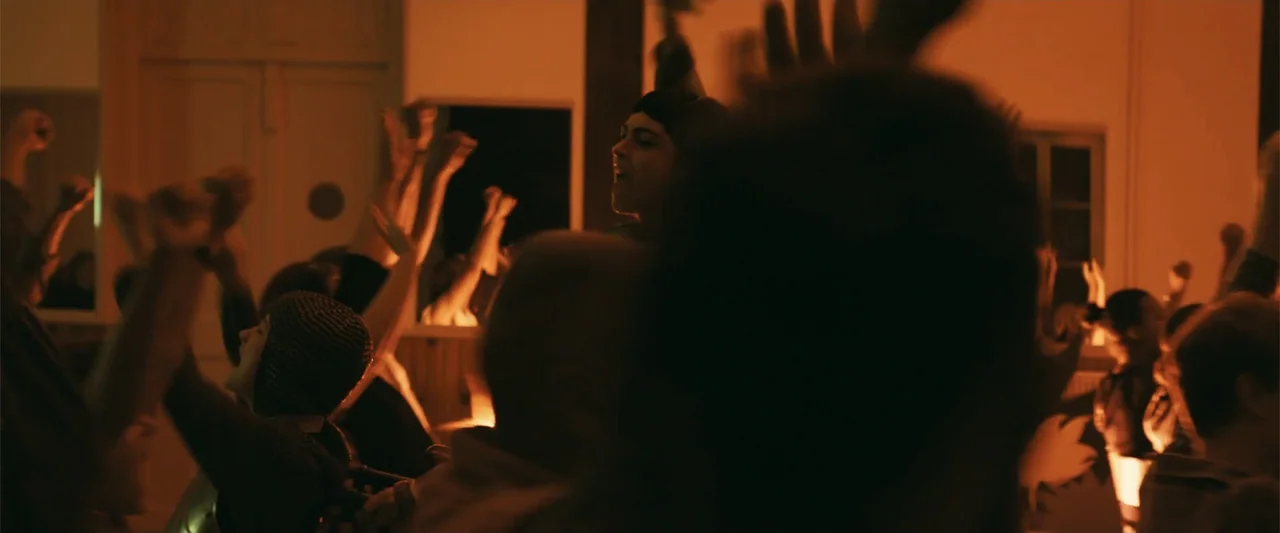 Dogman (2023) movie still: medium — A dimly lit indoor gathering space is crowded with people at night. Many individuals have their hands raised in…