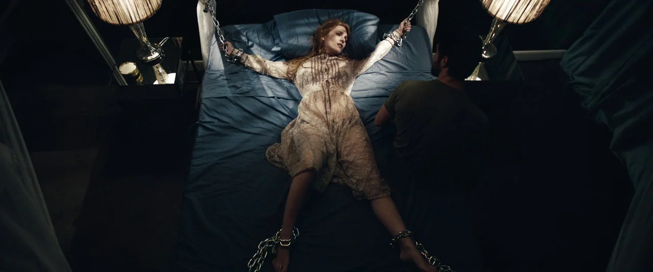 Kiss of the Damned (2012) movie still: fullbody — A dark bedroom with blue bedding features a bed with metal headboard posts. A woman lies chained…