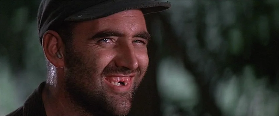 Deliverance (1972) movie still: closeup — A man stands outdoors with foliage visible in the out-of-focus background. The man looks toward the camera…