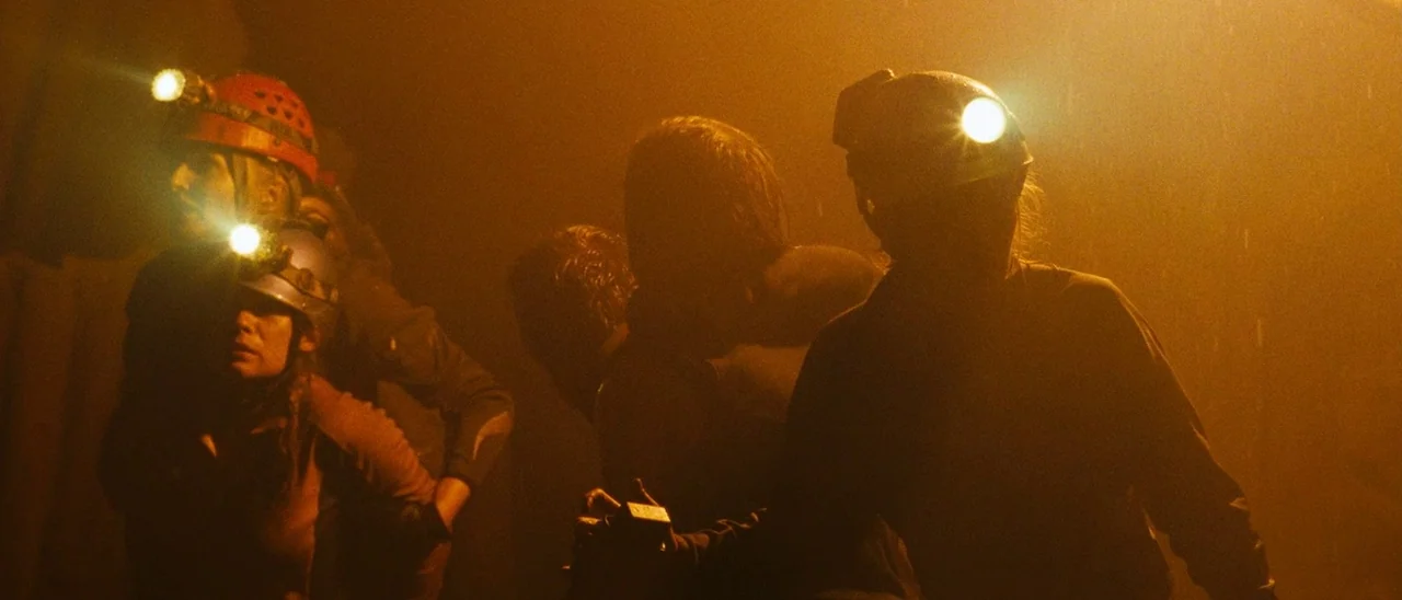 The Descent (2005) movie still: medium — A group of people navigate through a dark underground space illuminated by headlamps. Dust particles catch…