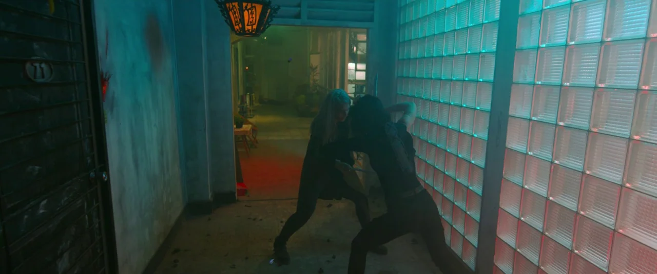 The Night Comes for Us (2018) movie still: fullbody — A dark corridor features a glass brick wall glowing with cyan light. Two women in dark…