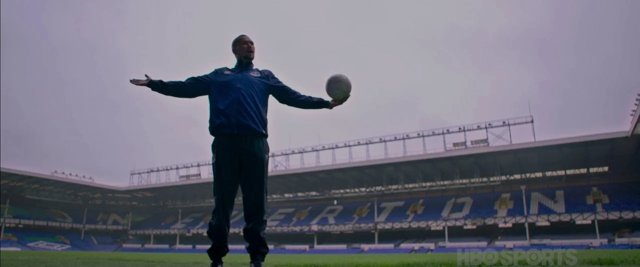 Creed (2015) movie still: fullbody — A vast, empty sports stadium features tiered blue seating arranged around a green pitch. A man stands at the…