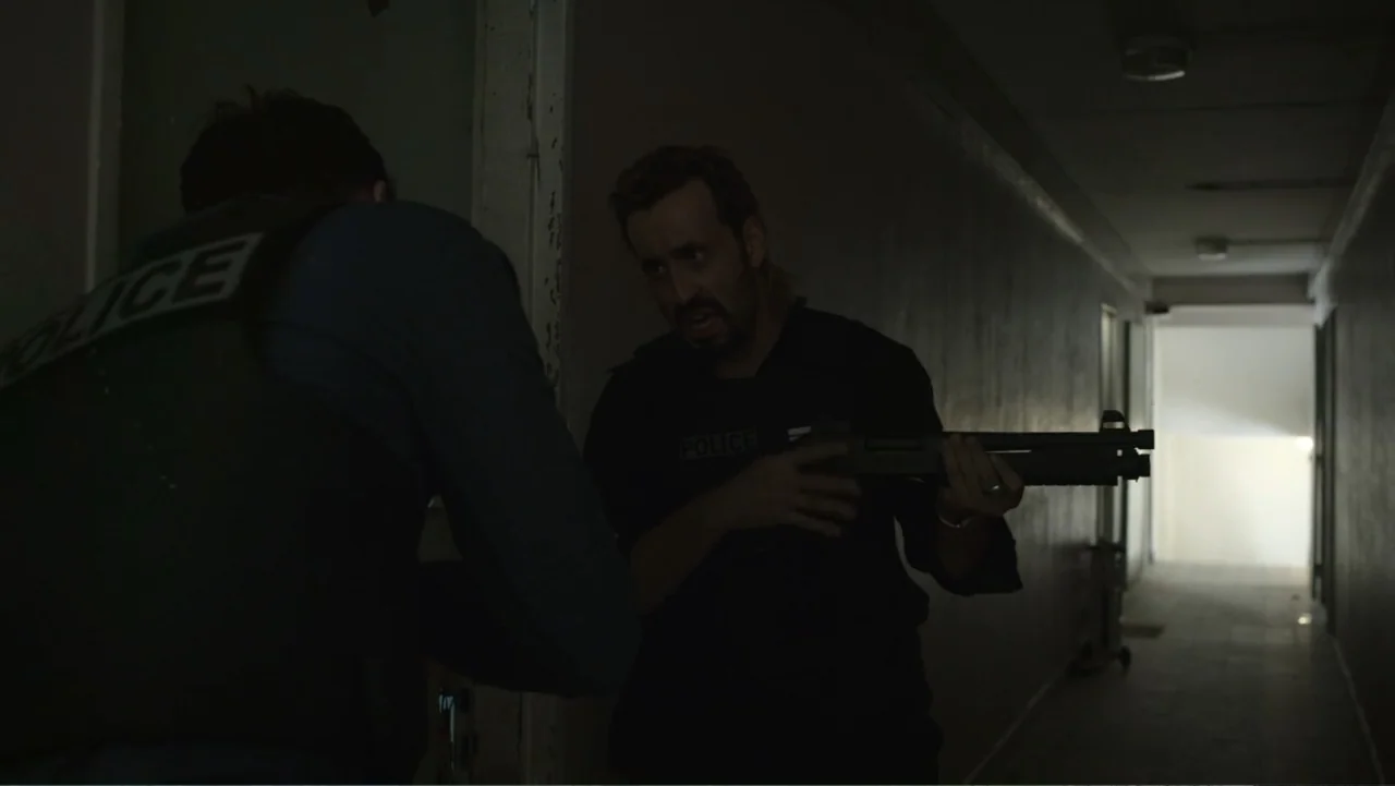 Unknown #1 movie still: medium — A dimly lit narrow apartment corridor features walls in shadow and a distant light source. A man in tactical gear…