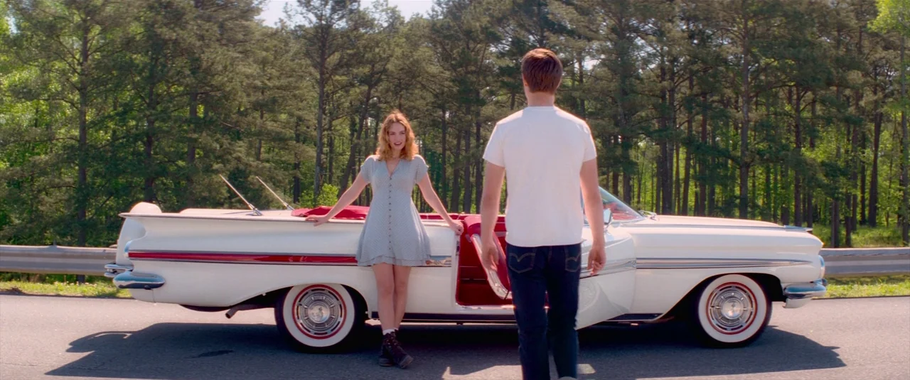 Baby Driver (2017) movie still: fullbody — A paved road runs alongside a dense forest of pine trees under daylight. A woman stands by a vintage…