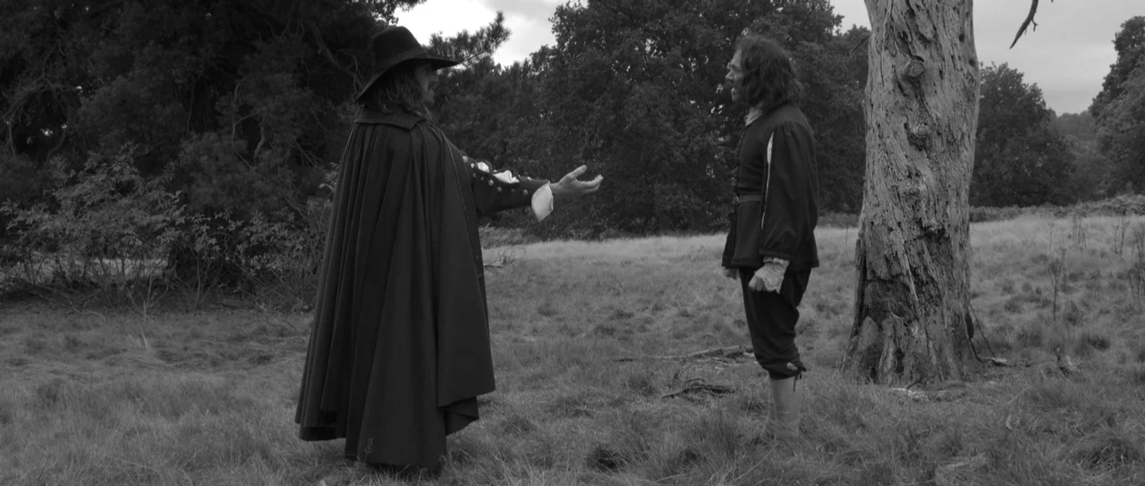 A Field In England (2013) movie still: fullbody — A desolate field features a large, gnarled bare tree on the right side. Two men dressed in…