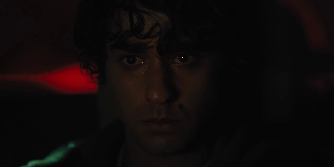 Hereditary (2018) movie still: closeup — The interior of a car at night is dark and mostly obscured. A young man with dark, curly hair looks…
