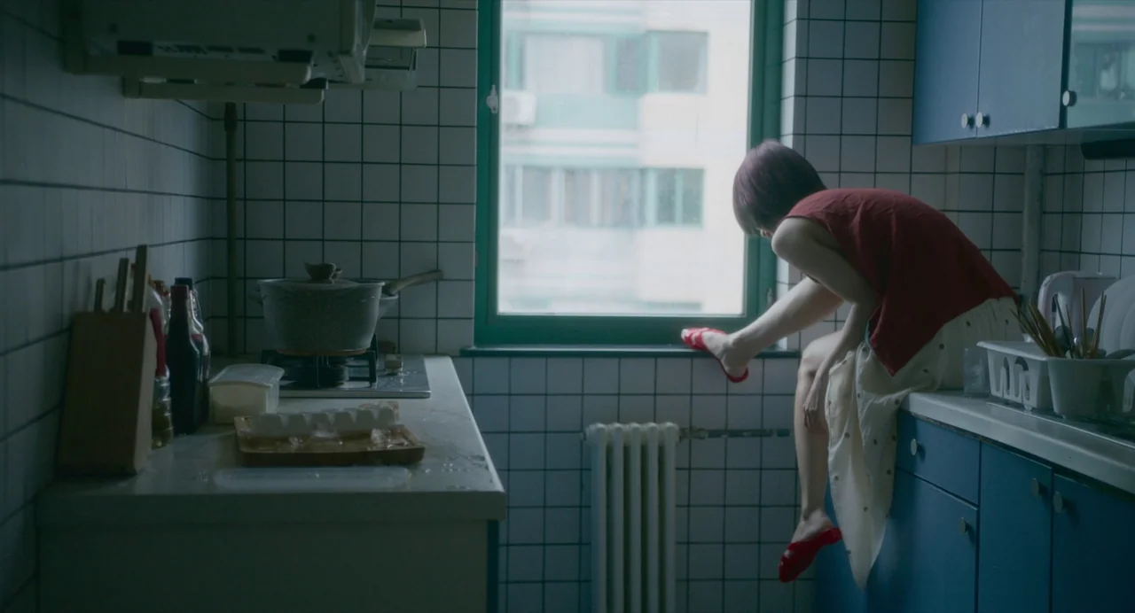 White River (2023) movie still: medium — A small kitchen features white tiled walls and blue cabinets with a centered window. A young woman sits…