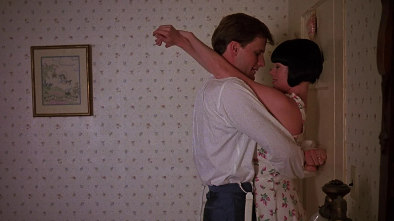 Something Wild (1986) movie still: medium — A room features floral-patterned wallpaper and a framed drawing hanging on the wall. A man and a woman…
