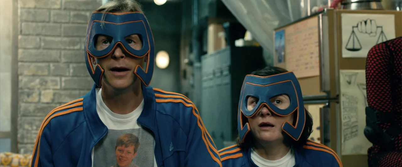 Kick-Ass 2 (2013) movie still: medium — A dimly lit basement room features brick walls and a refrigerator covered in notices. Two people in matching…