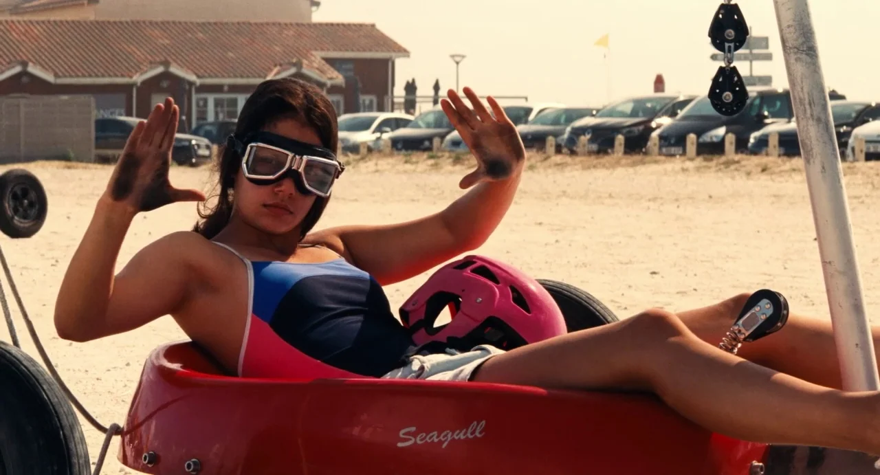 Ava (2017) movie still: medium — A sunny sandy coastal parking lot with cars parked in the distance. A woman sits in a red land yacht holding her…