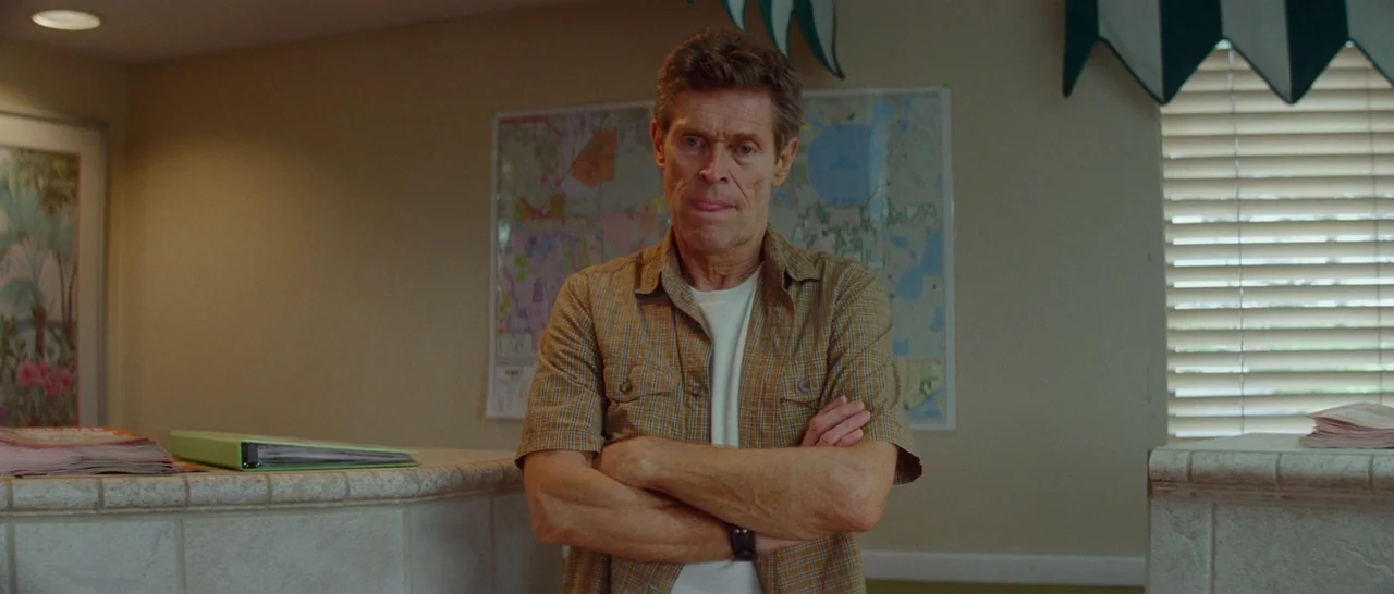 The Florida Project (2017) movie still: medium — A dated motel lobby features a tiled front desk and a map on the wall. A man stands behind the…