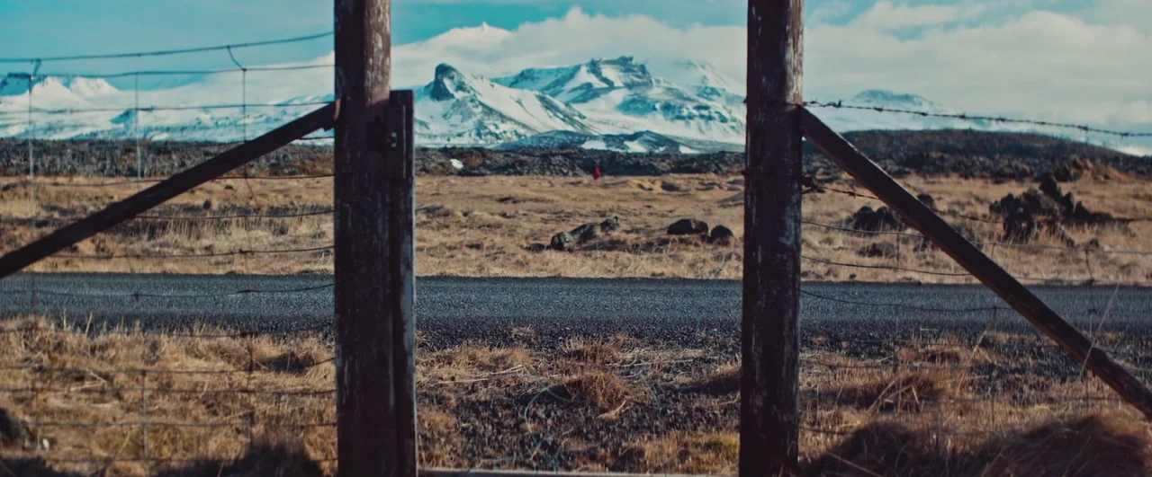 Rift (2017) movie still: wide — A rural asphalt road runs horizontally between two vertical wooden gate posts. Distant snow-capped mountains loom…