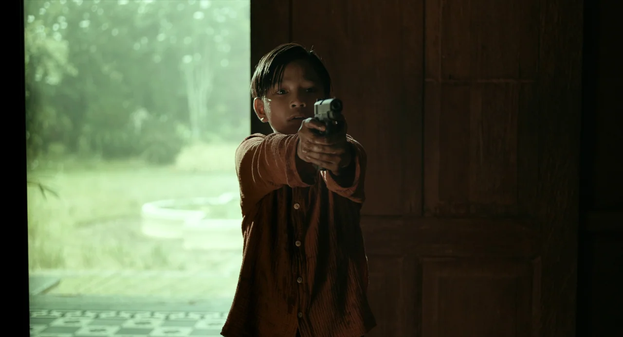 In My Mother’s Skin (2023) movie still: medium — A dark wooden room frames the bright view of an overgrown outdoor area. A child stands in the…