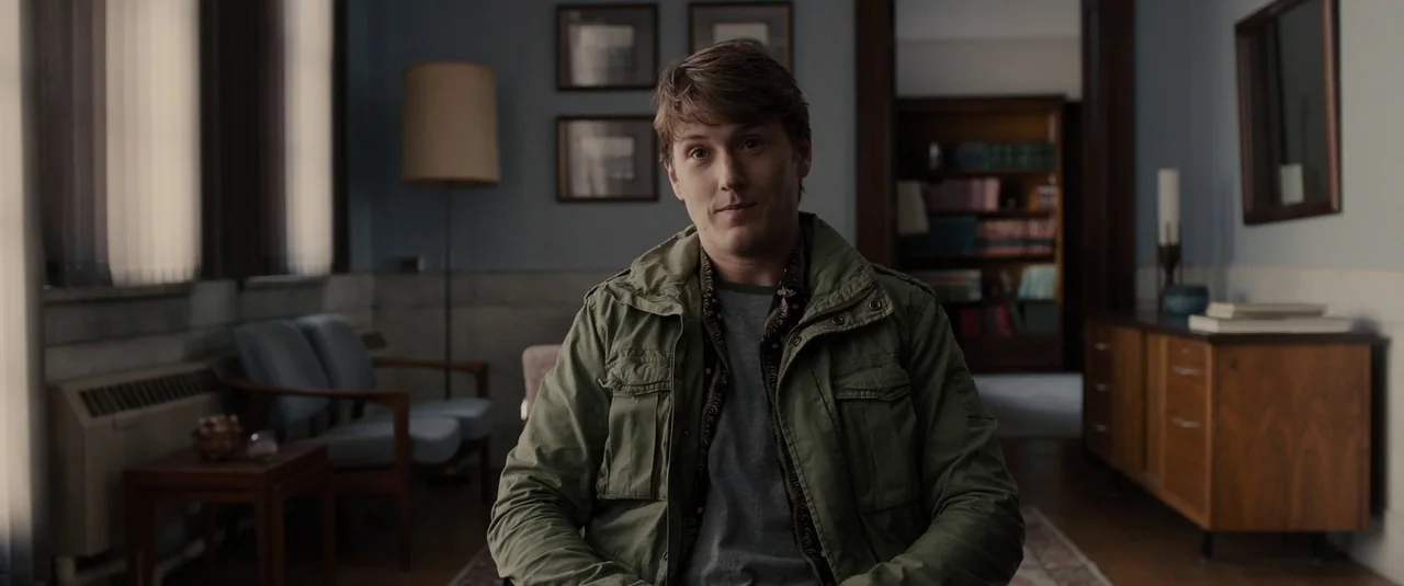 Glass (2019) movie still: medium — A quiet room features blue walls, framed artwork, and wooden furniture in the background. A young man sits in the…