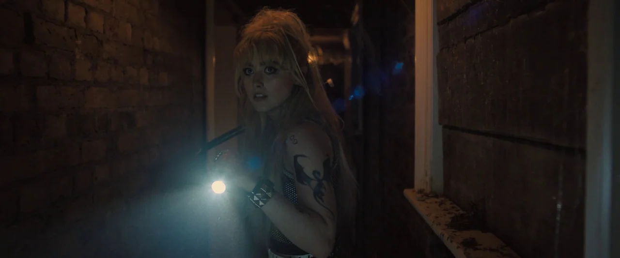 Abigail (2024) movie still: medium — A dark brick-lined corridor with a rough texture and dim surroundings. A young blonde woman shines a flashlight…