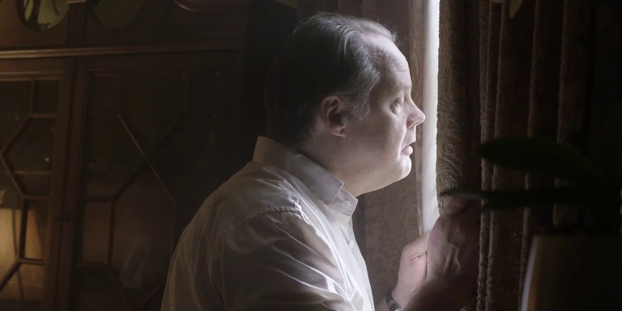Jakob’s Wife (2021) movie still: medium — A dark, dimly lit room contains a large wooden cabinet in the background. A man stands by a window,…