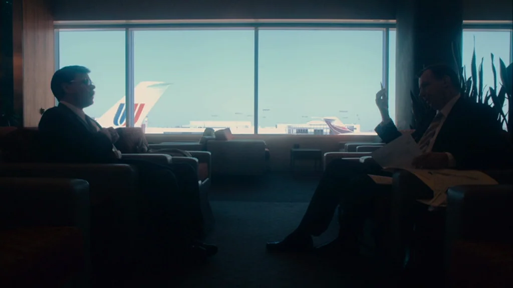 The Informant! (2009) movie still: wide — An airport lounge with modern armchairs and a wide window overlooking parked airplanes. Two businessmen in…