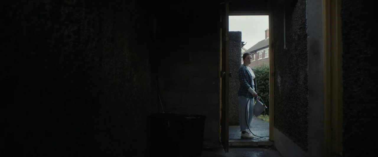 You Are Not My Mother (2021) movie still: fullbody — A woman stands in the doorway of a dark shed looking outward. She holds a garden hose and a…