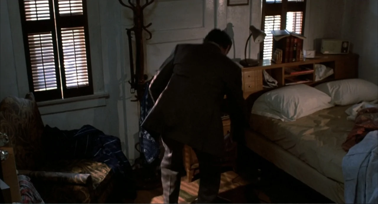 Murphy's Law (2009) movie still: medium — A cluttered bedroom with a made bed and patterned armchair near a window. A man in a suit bends down to…