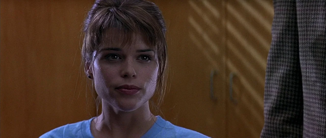 Scream (1996) movie still: closeup — An interior space with wood-paneled walls features a person positioned in the foreground. A young woman looks…