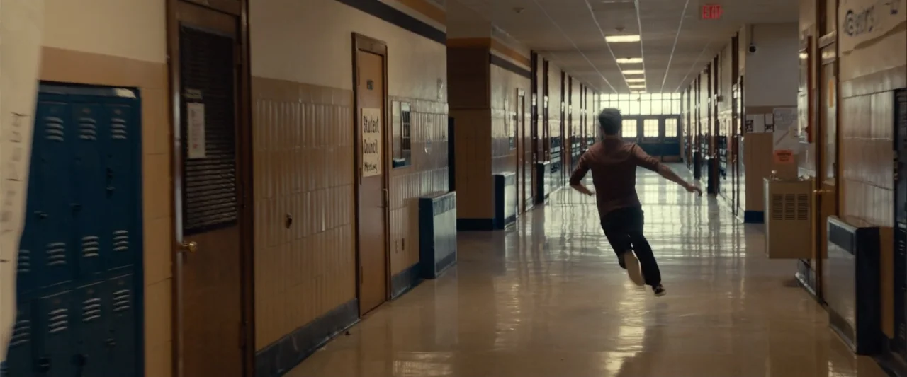 Frontera (2014) movie still: fullbody — A long school corridor with tan tiled walls and rows of blue metal lockers. A young man runs through the…