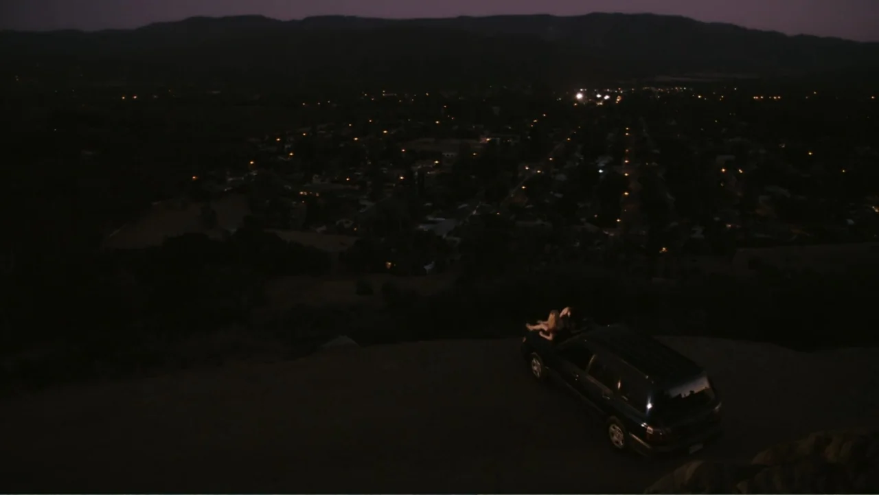 Easy A (2010) movie still: wide — A dark hilltop overlooks a sparsely lit residential town at night. Two people sit on the roof of a parked SUV on a…