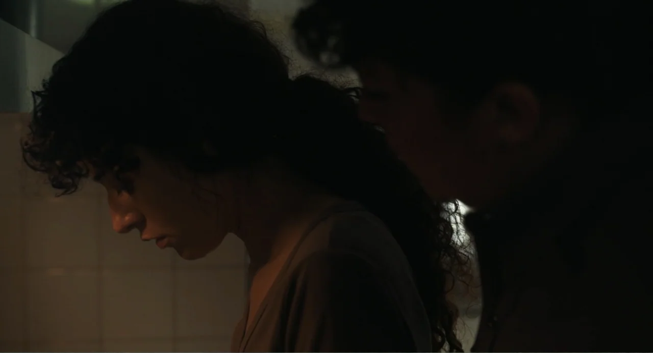 Belén (2025) movie still: closeup — A low-light bathroom interior features white tiled walls in the background. A woman with dark curly hair faces…