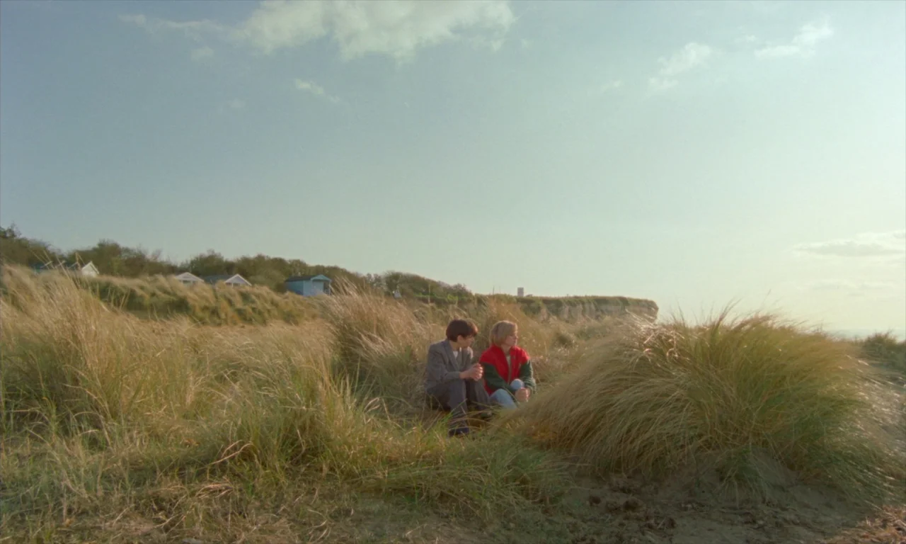 Spencer (2021) movie still: wide — A grassy coastal dune environment features distant small houses on a ridge. A boy and a girl sit together amongst…