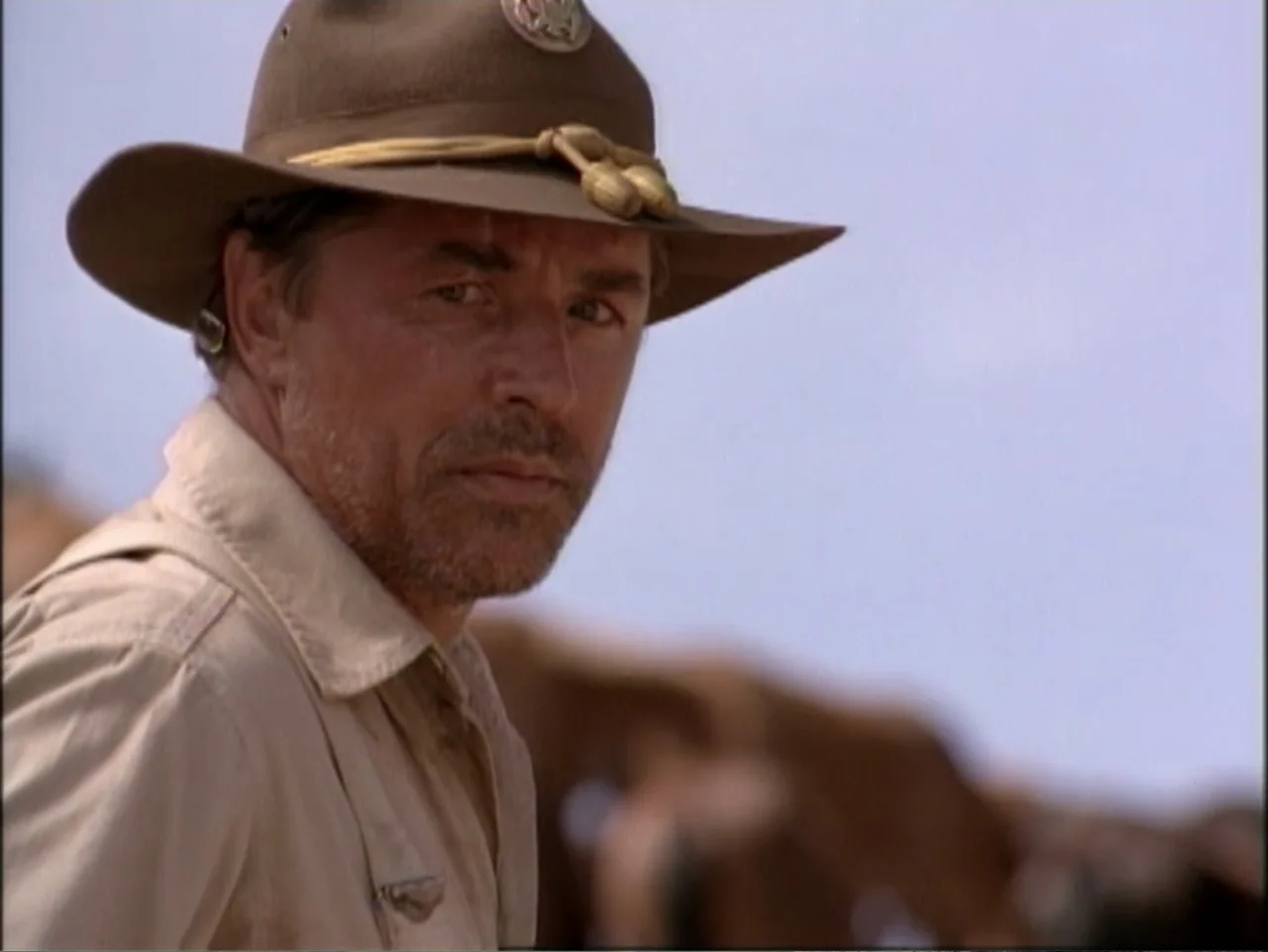 In Pursuit of Honor (1995) movie still: medium — An outdoor field setting under a bright, clear sky. A man in a military hat gazes forward while…