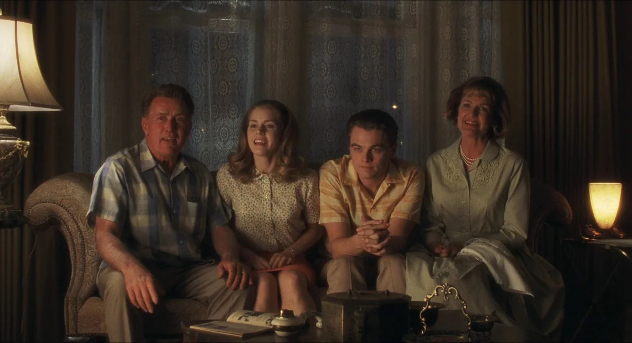 Catch Me If You Can (2002) movie still: medium — A 1960s suburban living room at night featuring patterned curtains and a sofa. Four people sit…