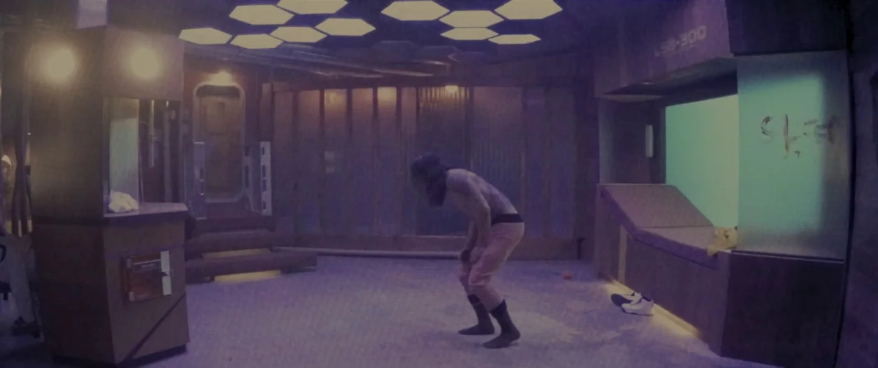 Domain (2017) movie still: fullbody — A futuristic holding cell features hexagonal ceiling lights and padded wall panels. The lone subject stands in…
