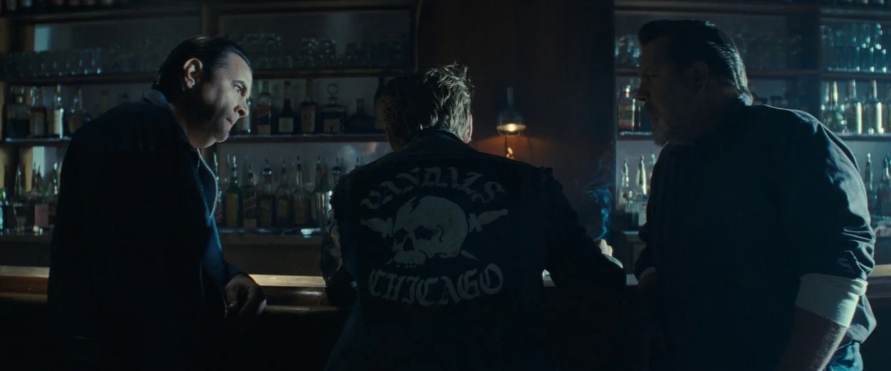 The Bikeriders (2023) movie still: medium — A dimly lit bar interior features liquor bottles lined up on dark wooden shelves. Three men sit along…