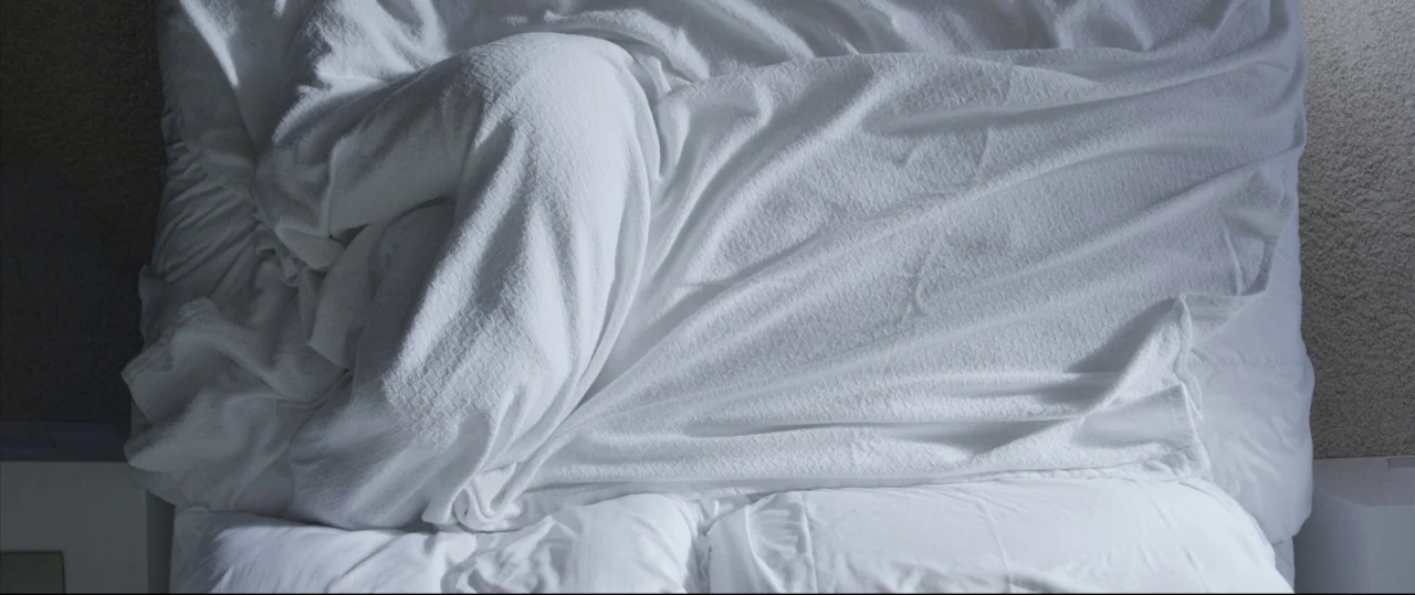 Sun Choke (2015) movie still: wide — An overhead view shows a large bed covered with rumpled white linens. Two pillows sit neatly at the base of the…