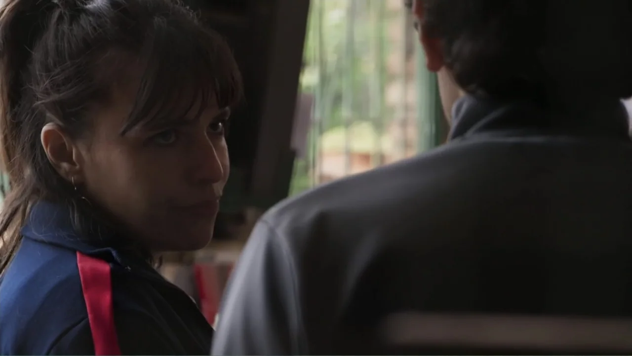 Smog in Your Heart (2022) movie still: medium — Two individuals sit near a window in a dimly lit indoor space. A woman in a navy athletic jacket…