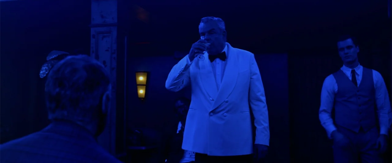Marlowe (2023) movie still: medium — A dimly lit room with dark wooden paneling and a glowing sconce light fixture. A man in a white dinner jacket…