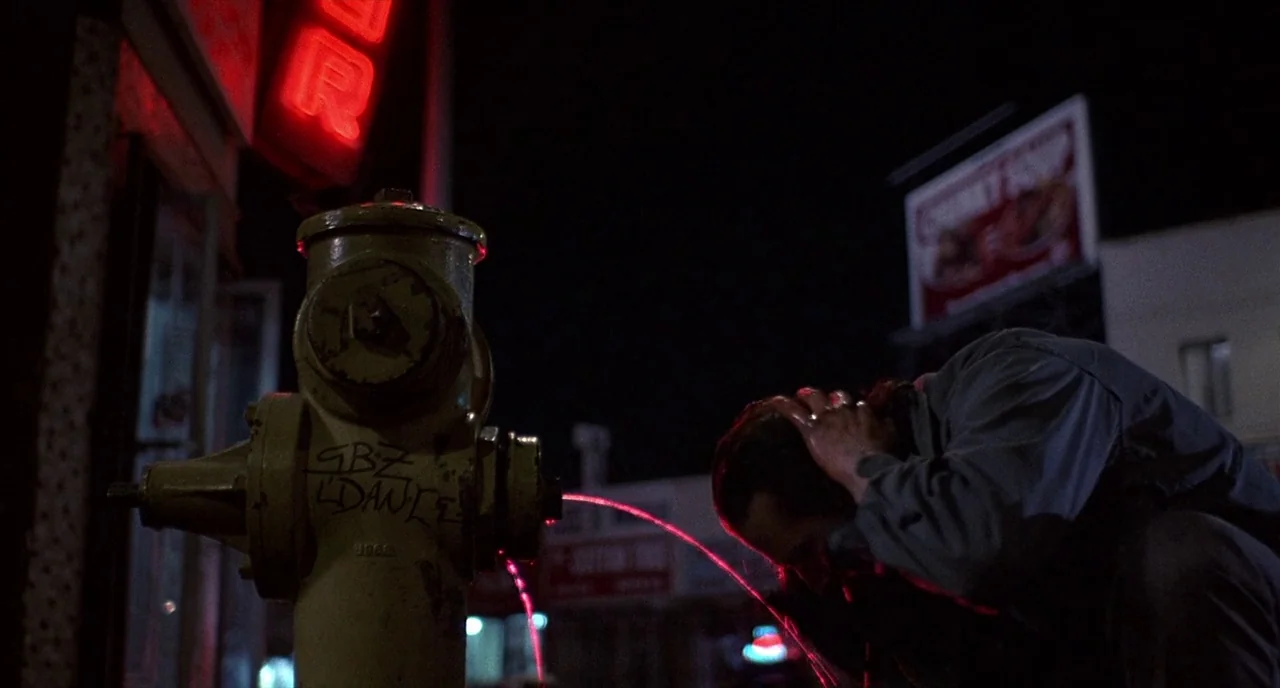 Barfly (1987) movie still: medium — A dark urban environment at night with a large yellow fire hydrant in focus. A man crouches beside the fire…