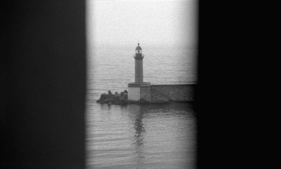 The Man From London (2007) movie still: wide — A stone lighthouse stands on a breakwater extending into a calm, grey sea. Thick black curtains frame…