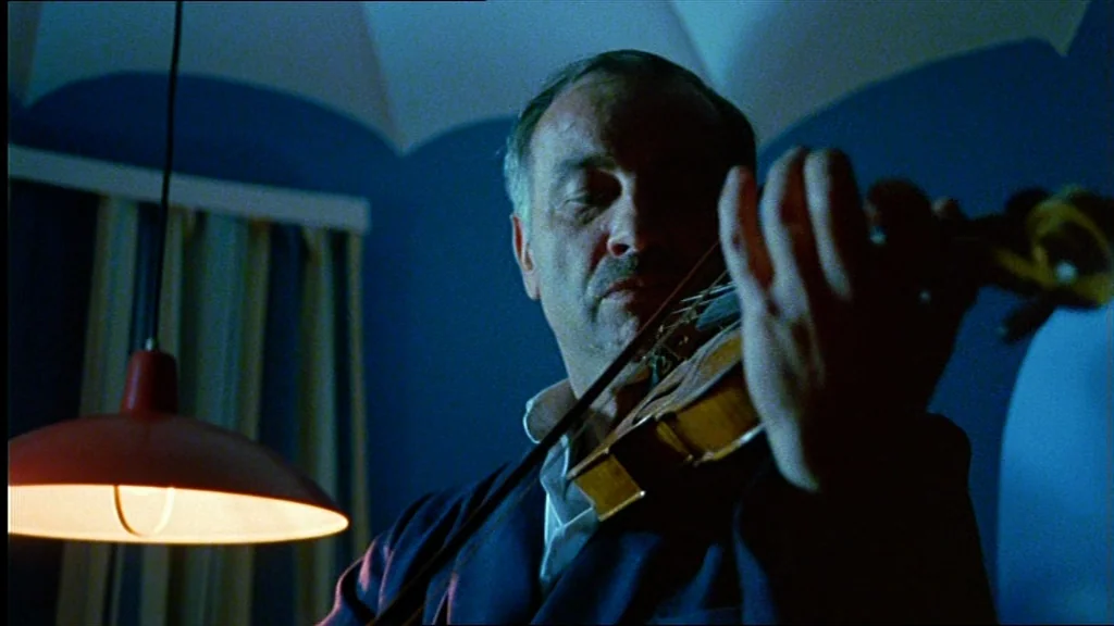 Lola (1981) movie still: medium — A man plays a violin in a room bathed in blue light. An orange lamp hangs prominently in the foreground beside him.
