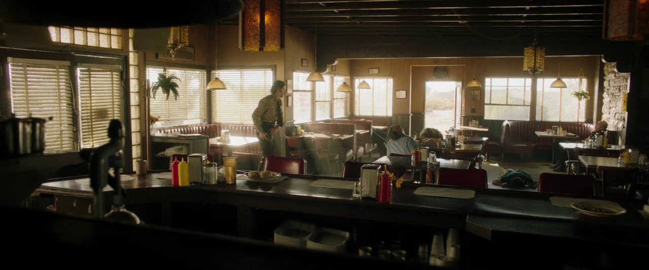The Last Stop in Yuma County (2023) movie still: wide — A quiet diner interior with red vinyl booths and dark wood tables under low ceilings. A man…