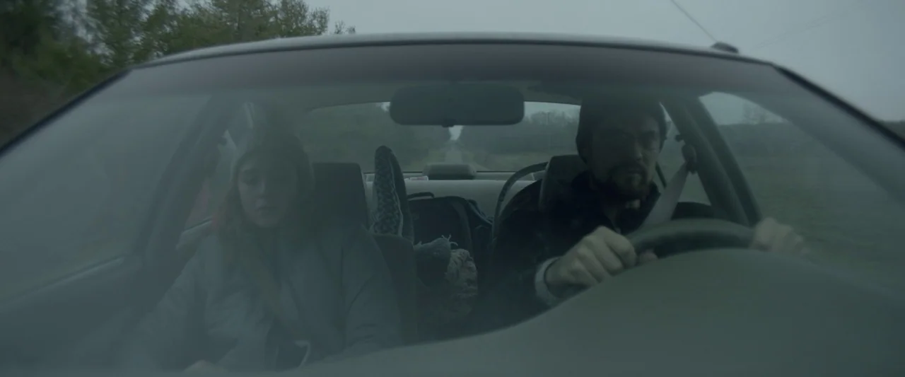 Here Alone movie still: medium — The interior of a moving car during an overcast, gray day on a country road. A man drives while a woman sits…
